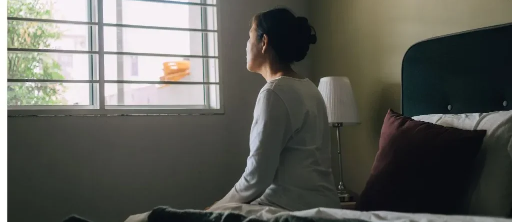 mulher sentada na cama triste olhando para a janela retratando morar sozinho e depressão